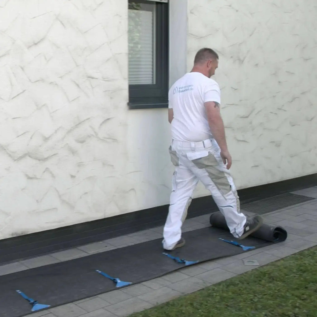 Ein mann in weißem t-shirt und beigen arbeitshosen rollt dunkelgraues belagsmaterial mit blauen griffen an der hermes auffangwanne 3.0 entlang einer strukturierten wand aus