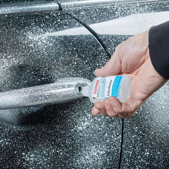 A clear plastic bottle with a white cap and blue sonax schlossentester label held by a hand for lock de-icing